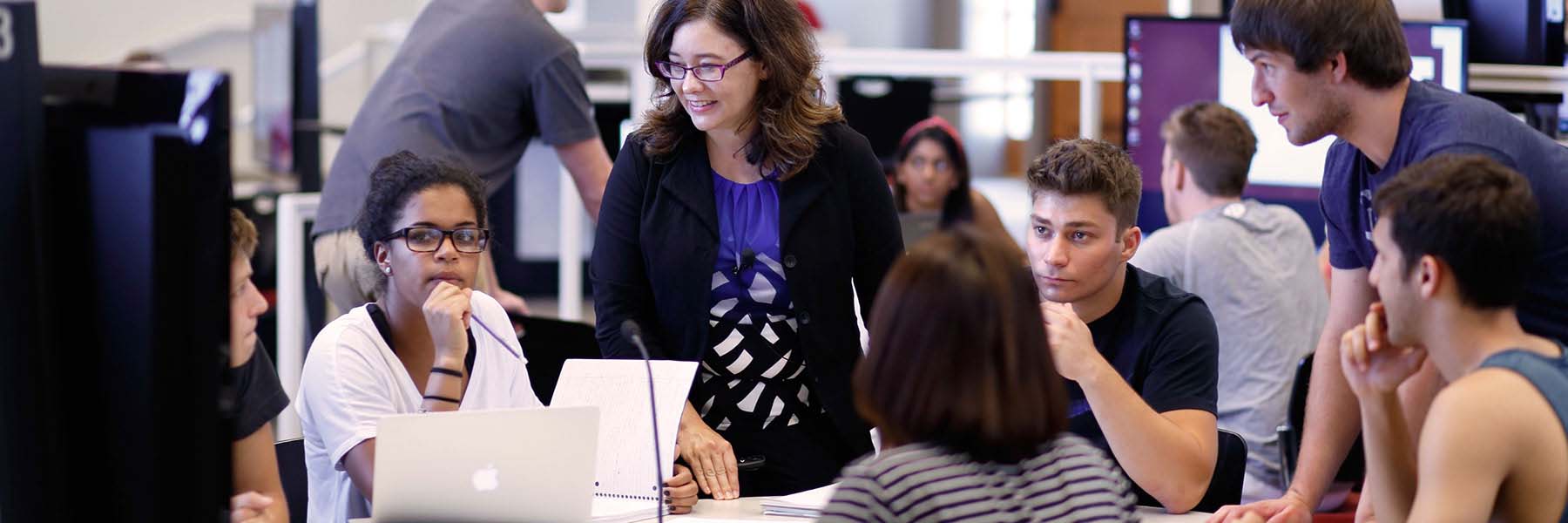 A professor talks with a group of students as they work on a project in a computer lab.