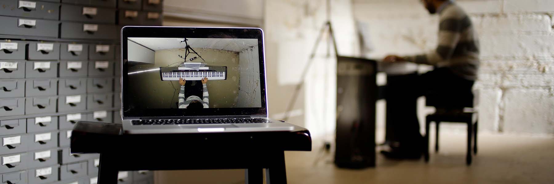 A computer screen in the foreground shows a student who is playing a piano in the background.